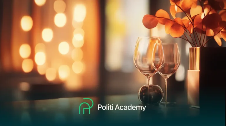 Duas taças de vinho vazias ao lado de folhas decorativas de laranja em uma mesa, com luzes quentes e desfocadas ao fundo. Logos e texto dizem "Politi Academy.