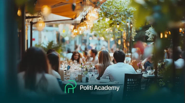 Pessoas jantando ao ar livre em um restaurante movimentado com luzes penduradas e vegetação. O logotipo da Politi Academy é visível na parte inferior.