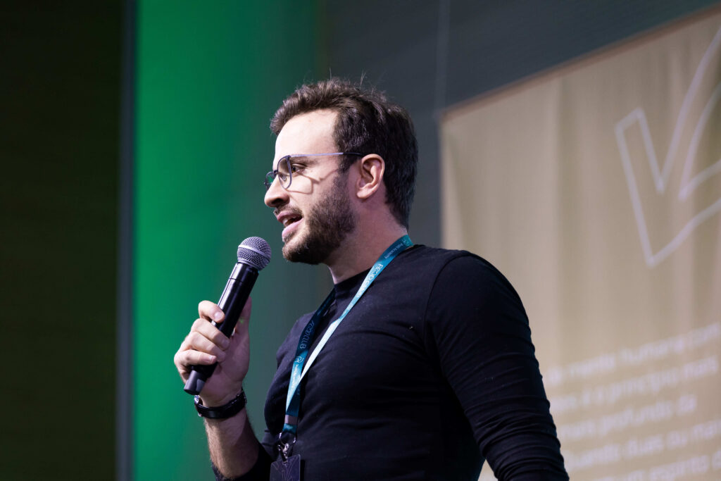 Um homem de óculos e camisa preta fala em um microfone no palco, com um cordão de Fincar Bandeira no pescoço e um fundo desfocado atrás dele.