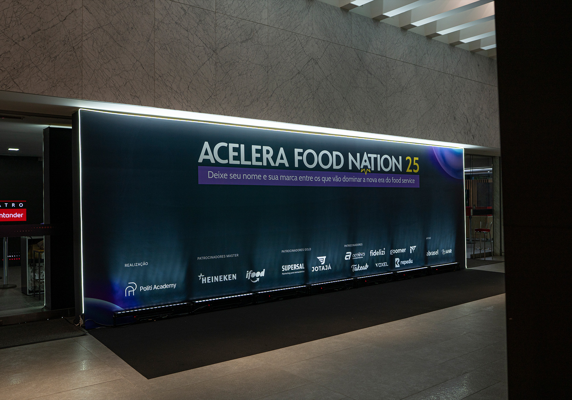 O grande banner do evento diz "ACELERA FOOD NATION 2.5", com logotipos de patrocinadores como Heineken, iFood e Santander, exibido em um local moderno e coberto - prepare-se para o Acelera Food Nation 2026 ou entre para a Lista de Espera!.