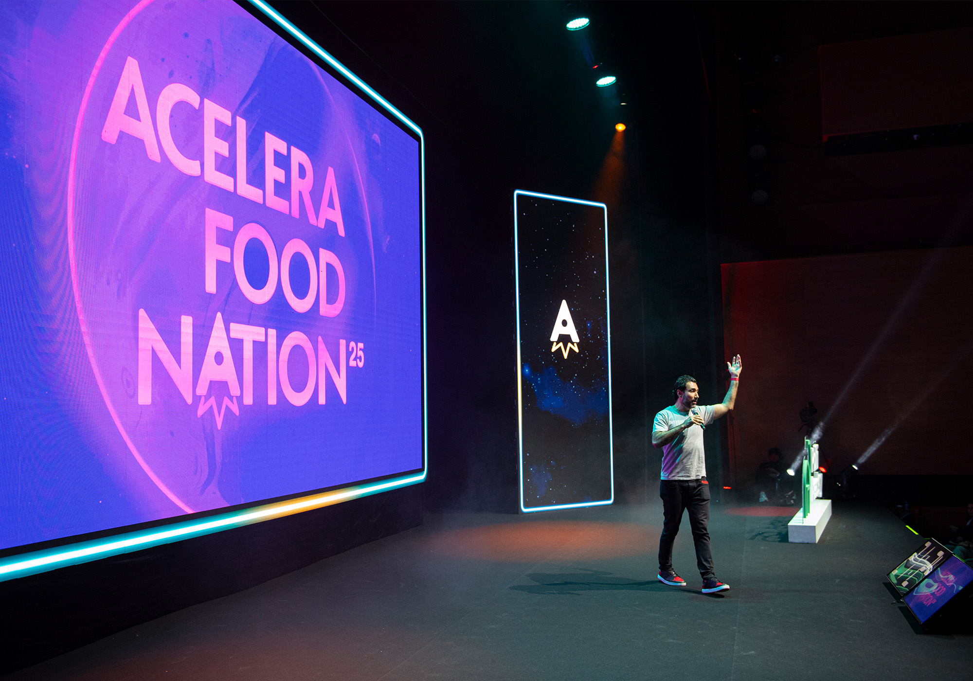 Um palestrante está no palco em frente a telas grandes que exibem "Acelera Food Nation 25" em um evento, enquanto os participantes aguardam ansiosamente para entrar na Lista de Espera para a esperada edição de 2026.
