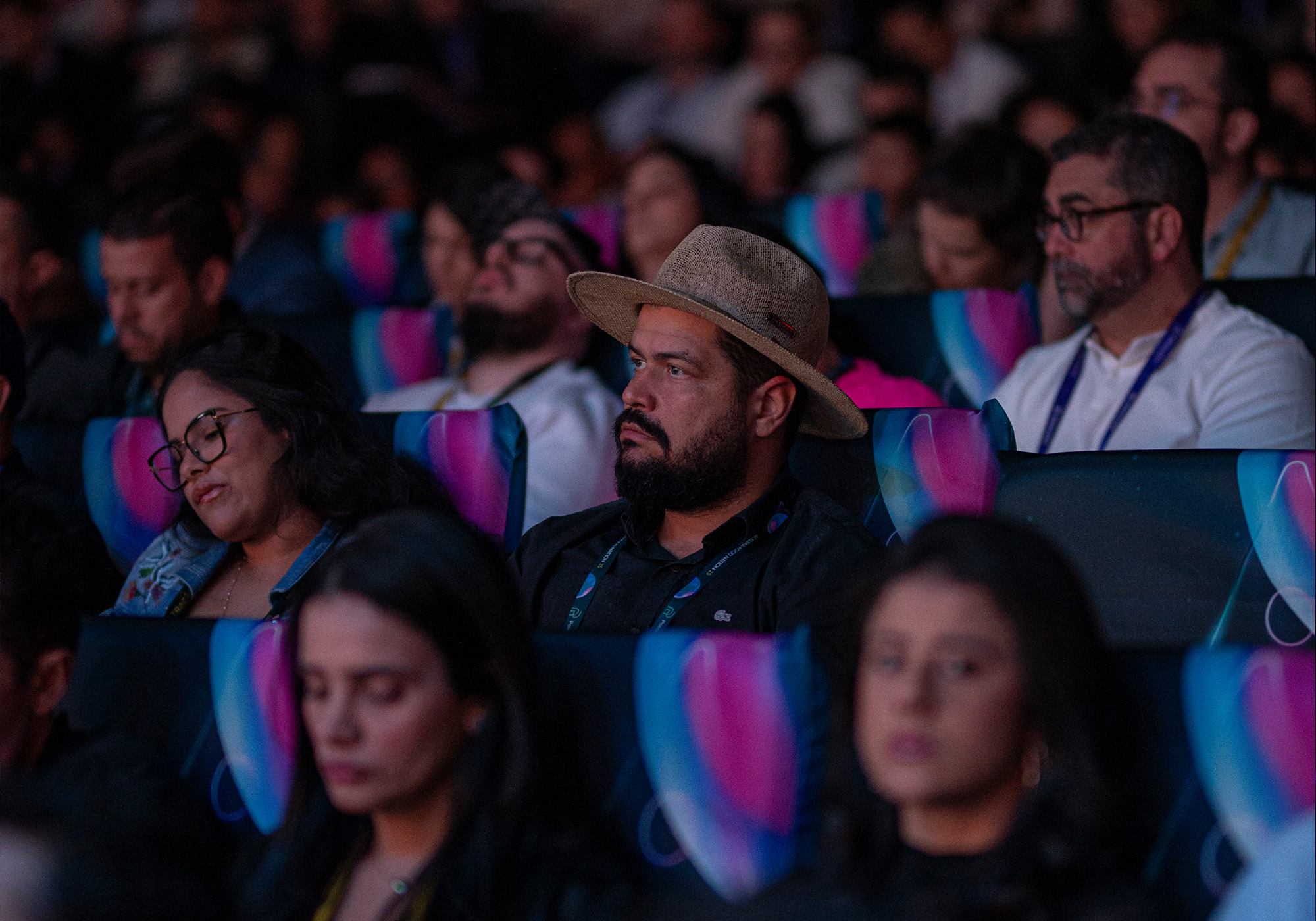 Os membros da plateia sentam-se em fileiras de assentos em estilo de teatro, assistindo atentamente a um evento; um homem no centro, talvez aguardando sua vez na Lista de Espera para o Acelera Food Nation 2026, usa um chapéu de abas largas.