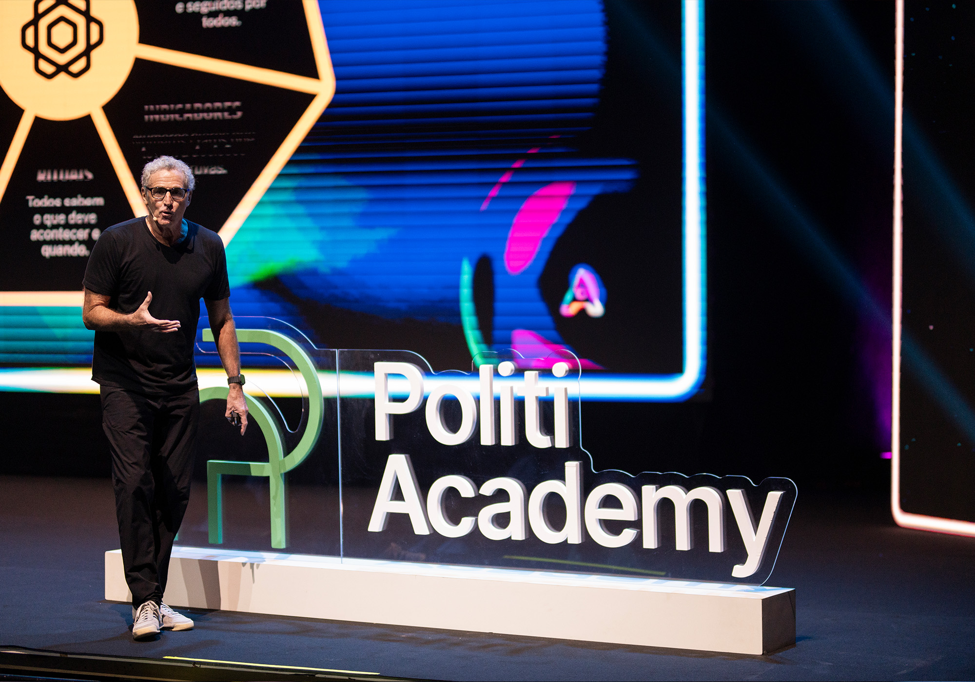 Um homem de roupa preta está no palco ao lado de uma placa que diz "Politi Academy", com uma tela digital grande e colorida ao fundo exibindo "Acelera Food Nation 2026".