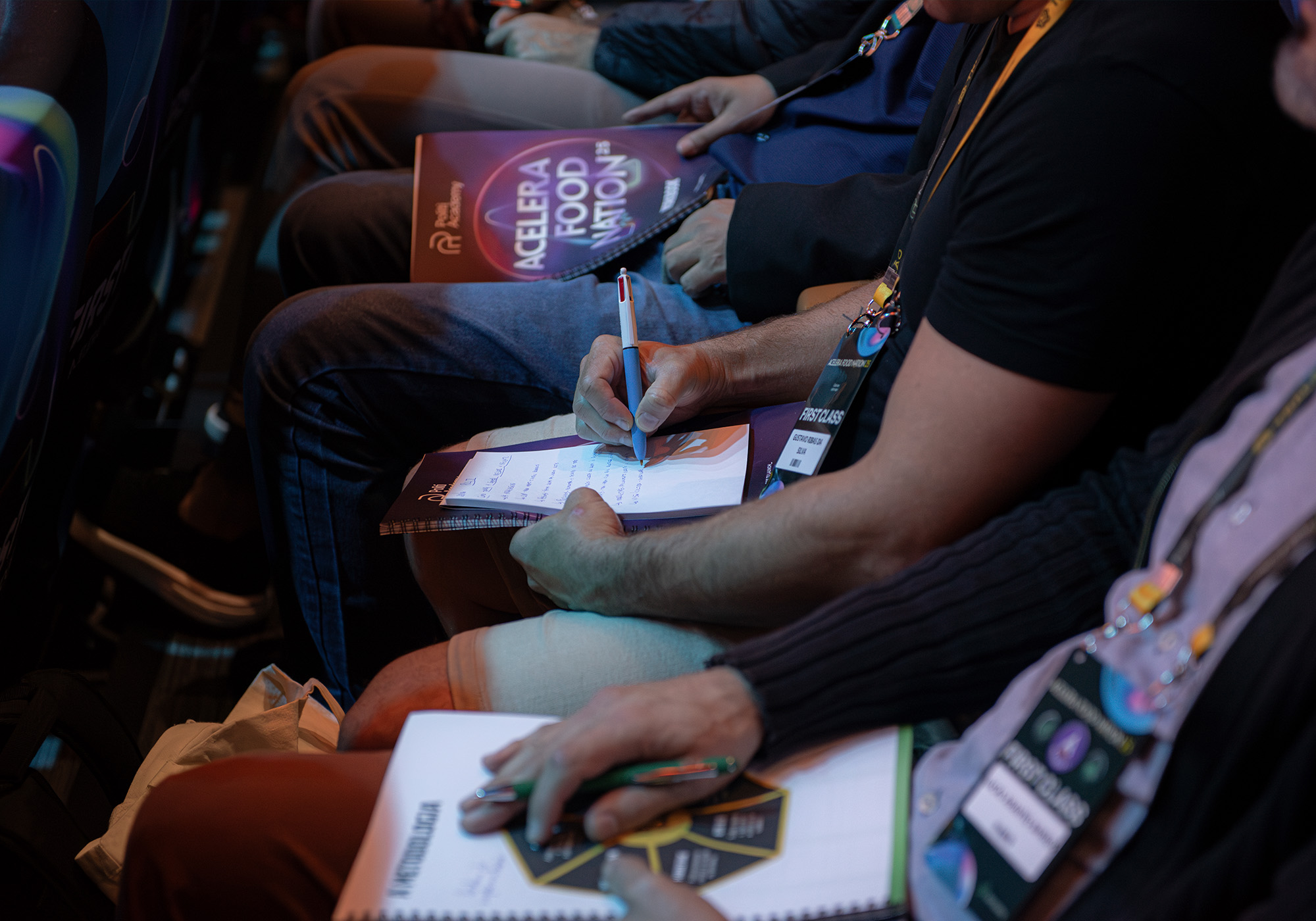 Várias pessoas sentadas em uma fileira fazem anotações em cadernos durante o evento Acelera Food Nation, usando cordões e segurando materiais do evento.