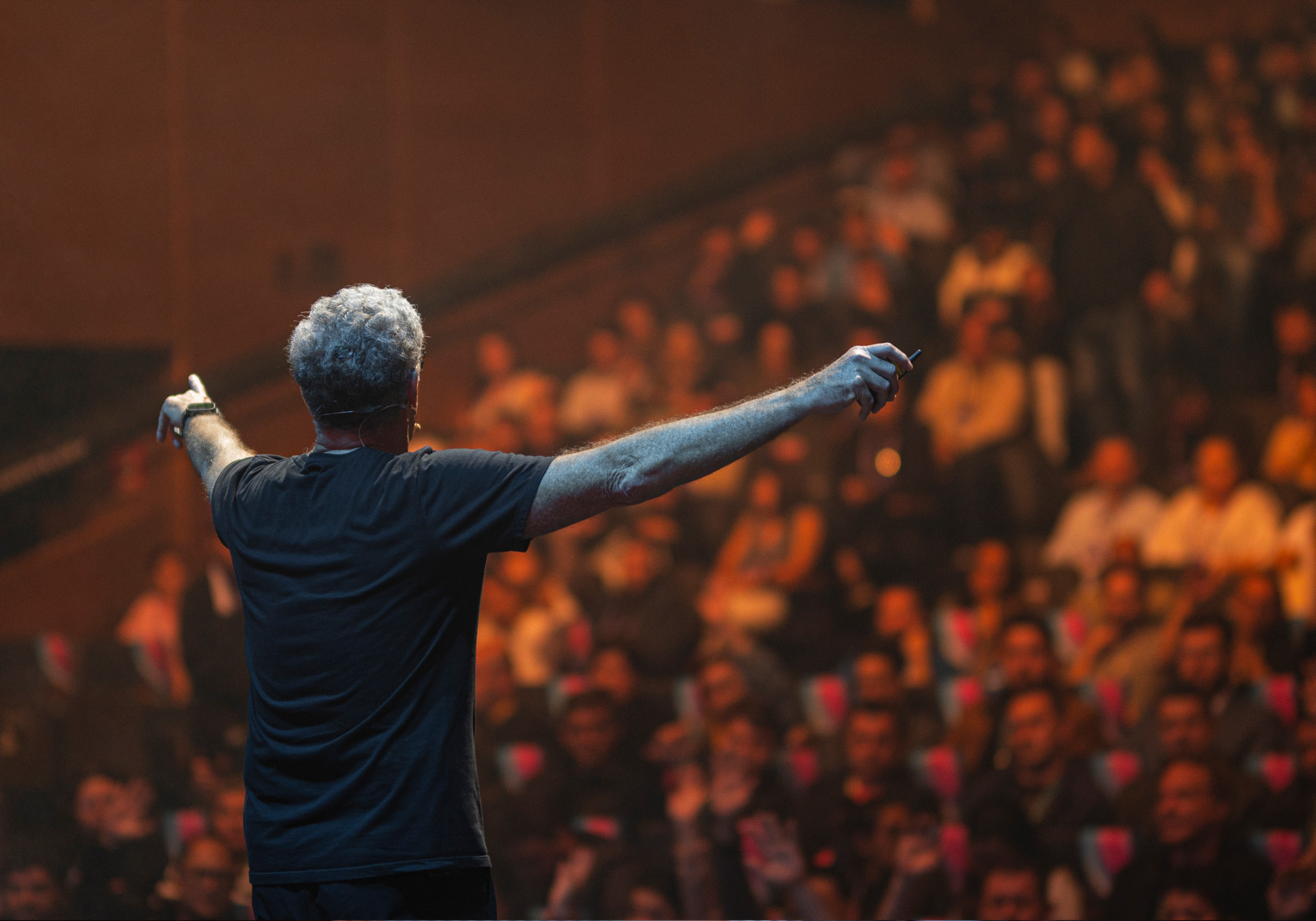 Um homem está no palco, de frente para uma plateia sentada, com os braços estendidos, em um auditório grande e pouco iluminado no evento Acelera Food Nation 2026.