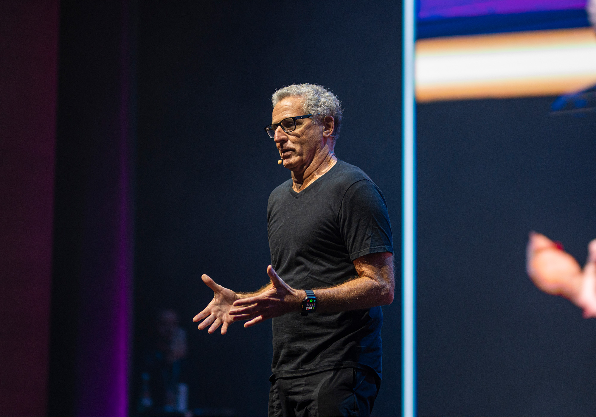 Um homem usando óculos, camiseta preta e um fone de ouvido com microfone fala no palco do evento Acelera Food Nation, gesticulando com as mãos abertas em frente a um fundo escuro, possivelmente discutindo a Lista de Espera de 2026.