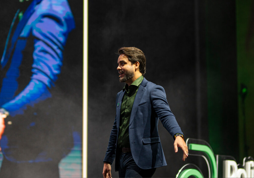 Um homem de terno azul e camisa verde entra no palco, com uma tela grande exibindo sua imagem ao fundo no Acelera Food Nation 2026. O público aguarda ansiosamente notícias sobre a lista de espera para as próximas inovações.