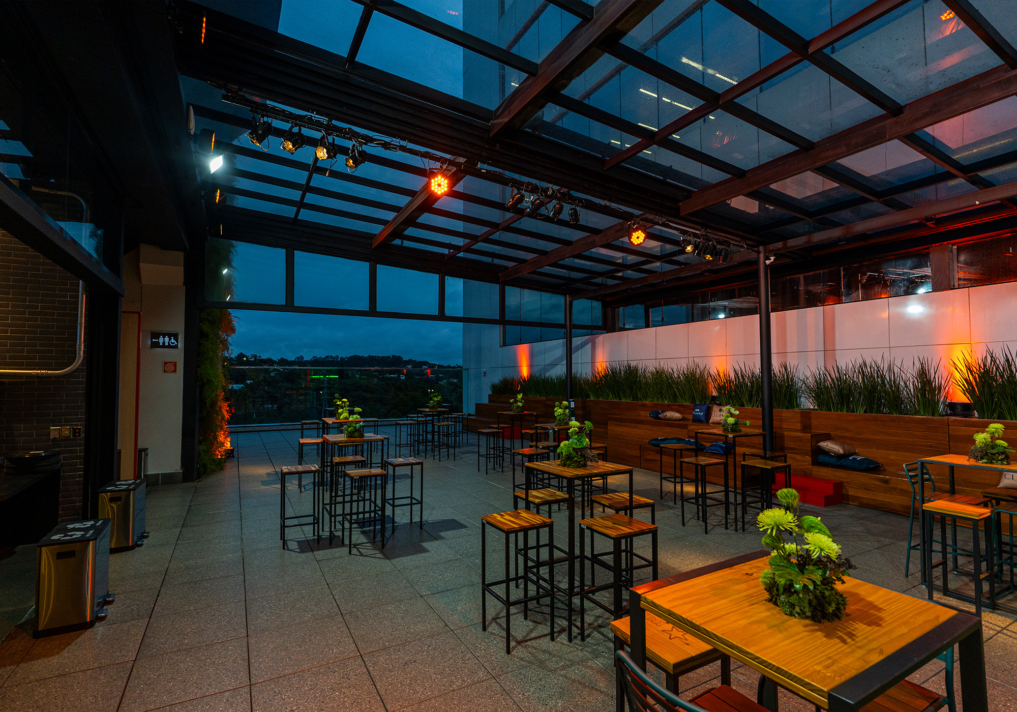 Bar na cobertura com teto de vidro, mesas e cadeiras altas, plantas verdes como centro de mesa e iluminação quente à noite - perfeito para curtir as vibrações do Acelera Food Nation. Junte-se à Lista de Espera 2026 para uma experiência exclusiva.