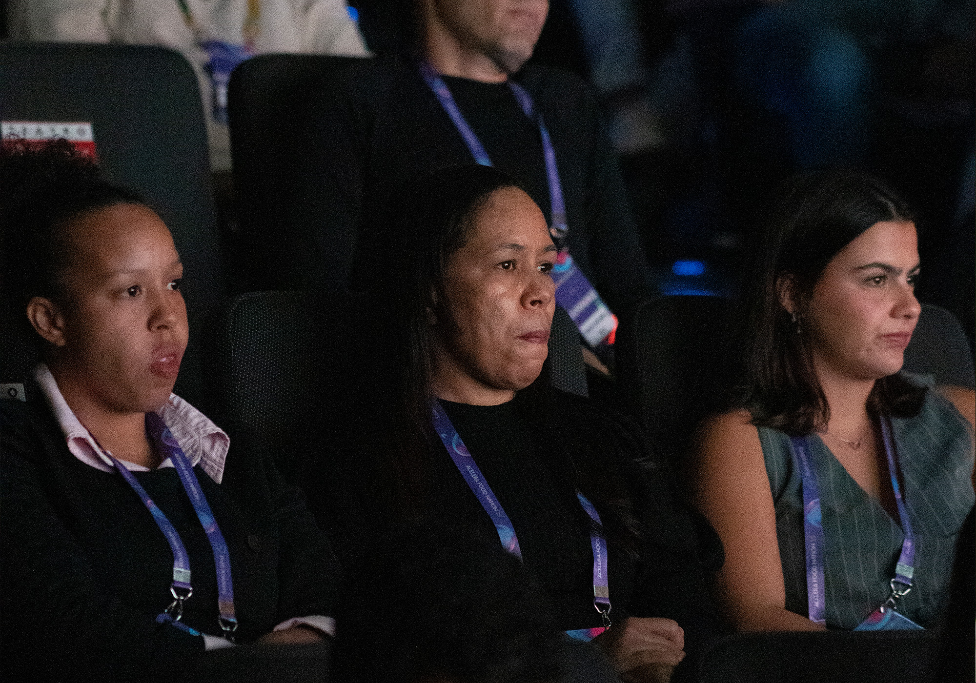 Três mulheres sentadas na plateia, usando cordões, observam atentamente algo fora da câmera no evento Acelera Food Nation, em um ambiente pouco iluminado.