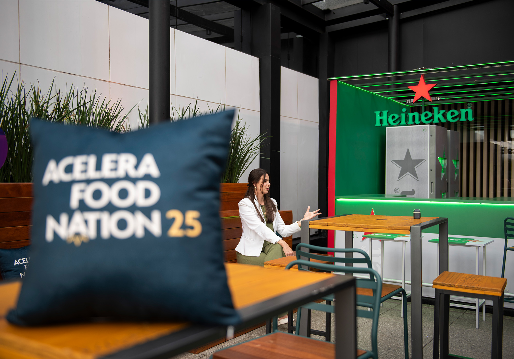 Uma mulher está sentada em uma mesa em uma área externa com sinalização da Heineken ao fundo; uma almofada em primeiro plano diz "Acelera Food Nation 25", sugerindo uma Lista de Espera exclusiva para esse evento especial.