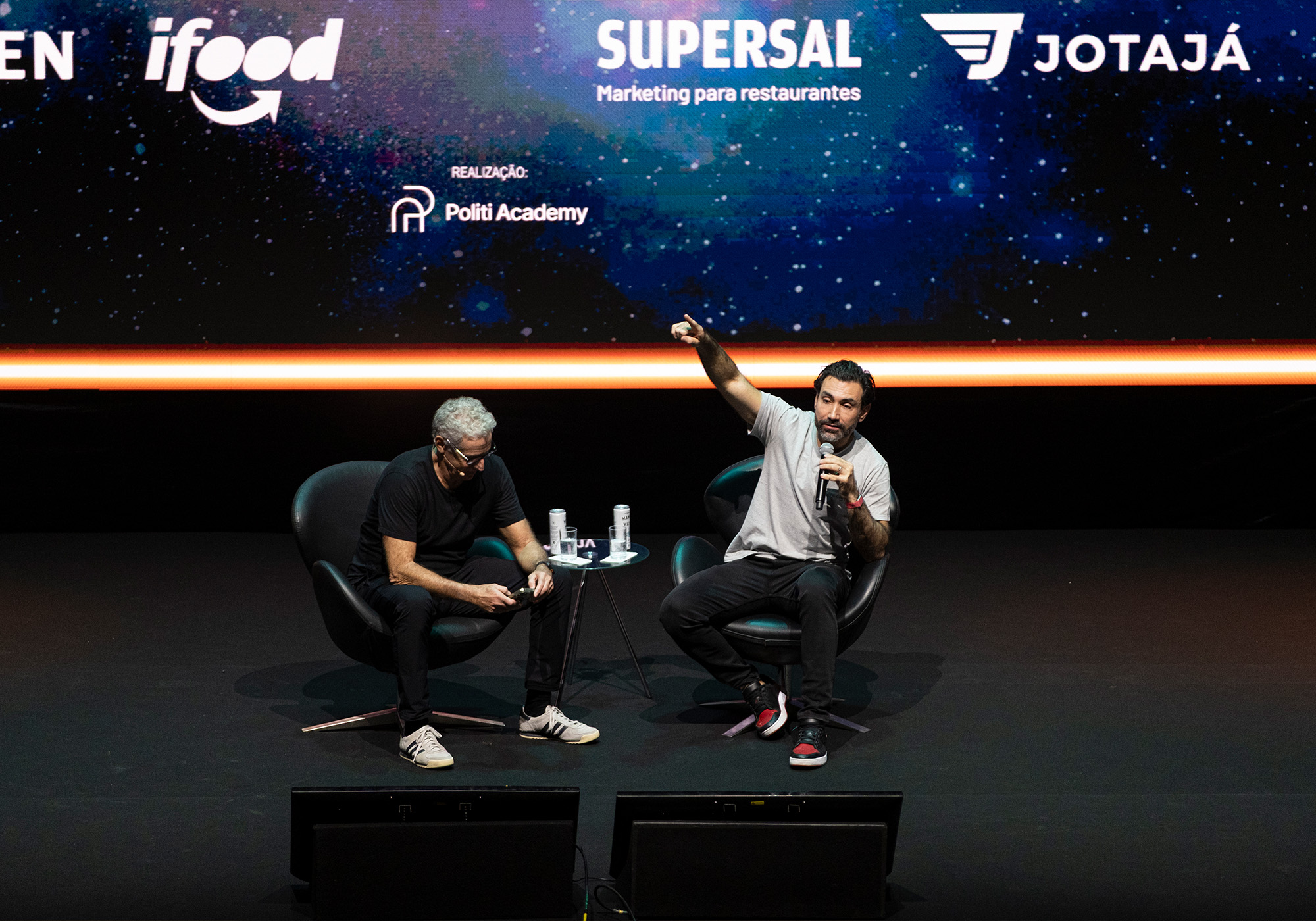 Dois homens sentados no palco com microfones durante um painel de discussão no Acelera Food Nation, com logotipos de patrocinadores exibidos em uma tela atrás deles. Um dos homens gesticula enquanto fala, pois o evento prevê o ano de 2026.