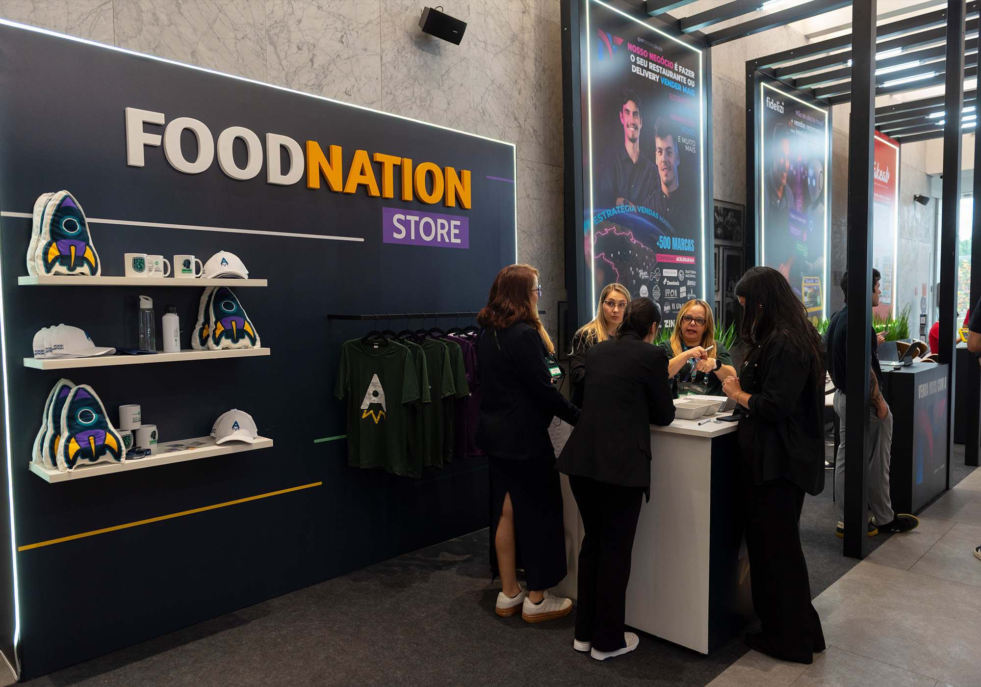 As pessoas ficam no balcão da FoodNation Store, cercadas por produtos da marca, como camisetas, bonés, canecas e brinquedos de pelúcia. Entre na Lista de Espera para obter itens exclusivos do 2026 Acelera Food Nation antes de todo mundo!.