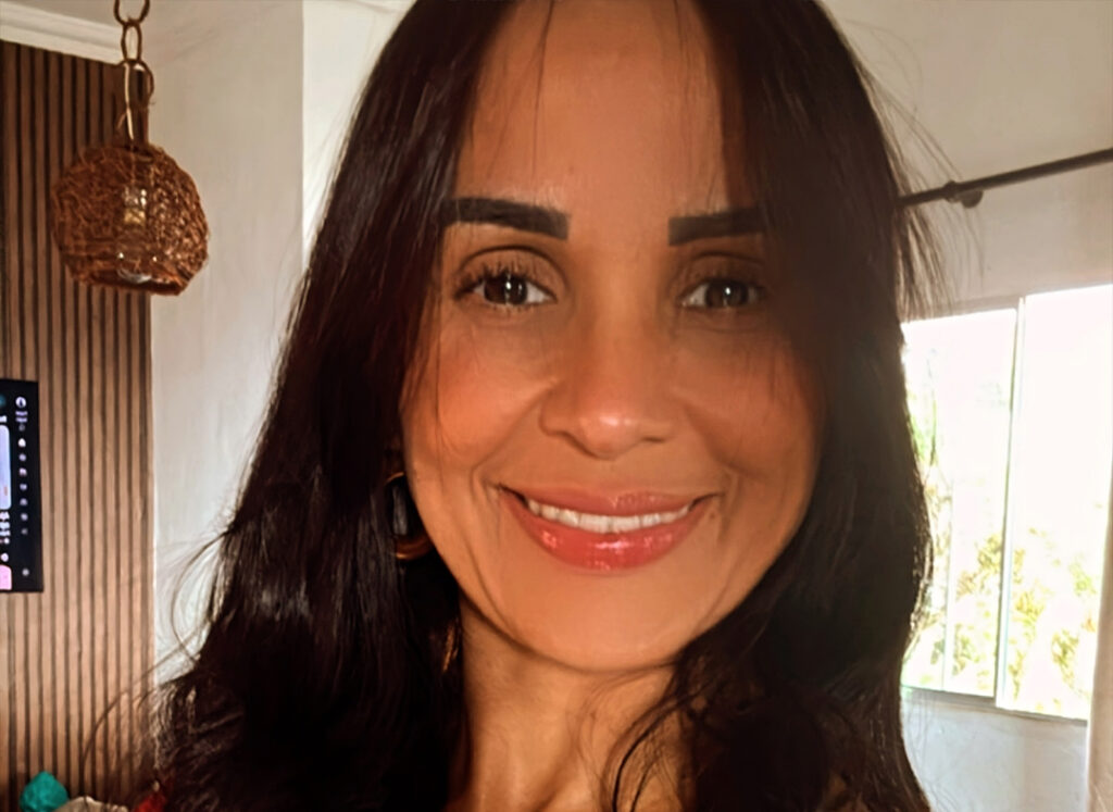 Mulher com longos cabelos escuros sorrindo em um ambiente interno, com uma janela e uma lâmpada de parede visíveis ao fundo, possivelmente na Fincar Bandeira após receber um diagnóstico positivo.