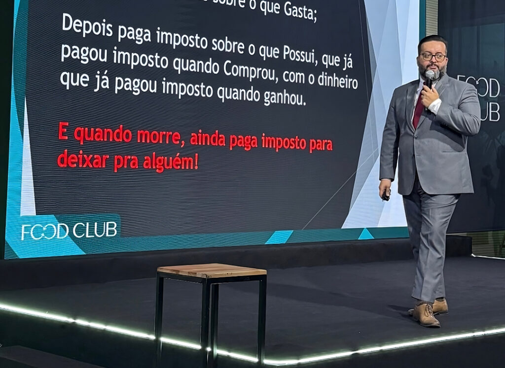 Um homem de terno cinza fala no palco ao lado de uma tela que exibe uma apresentação fiscal em português, com o texto Diagnóstico destacado em vermelho e o logotipo "FOOD CLUB".