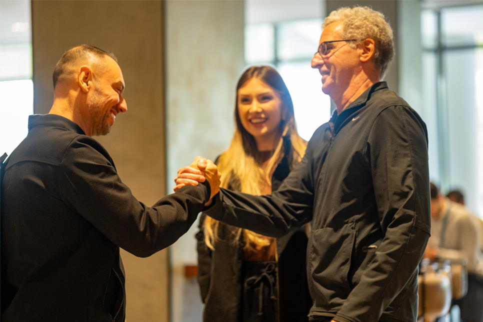 Dois homens se cumprimentam com um aperto de mão enquanto uma mulher fica entre eles, todos os três sorrindo em um ambiente interno bem iluminado.