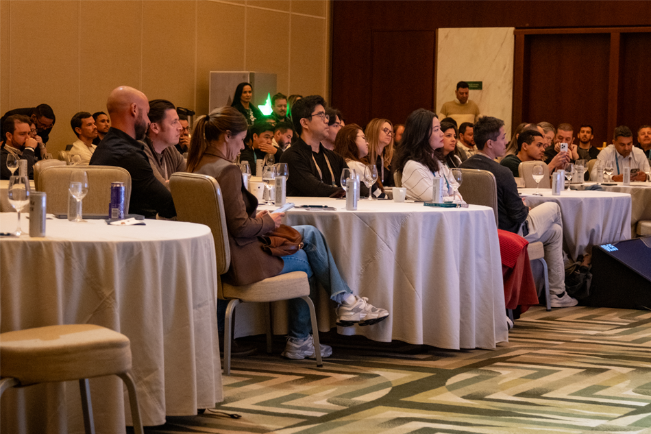 Um grupo de pessoas se senta em mesas redondas em uma sala de conferências, ouvindo atentamente uma apresentação.