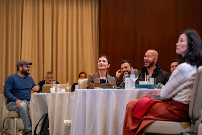 Um grupo de pessoas sentadas ao redor de mesas em uma sala de conferências, ouvindo atentamente e sorrindo durante um evento.