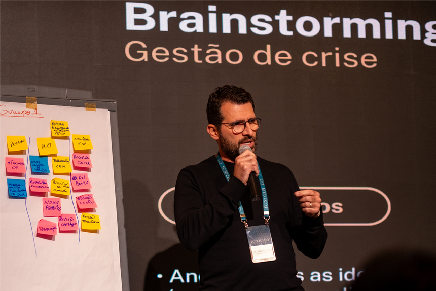 Um homem fala em um microfone durante uma apresentação sobre gerenciamento de crises, com um quadro de notas adesivas coloridas ao seu lado e "Brainstorming Gestão de crise" exibido ao fundo.