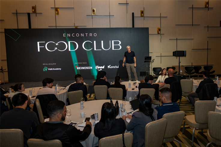 Um palestrante no palco se dirige aos participantes sentados em mesas redondas durante o evento Food Club em uma sala de conferências.