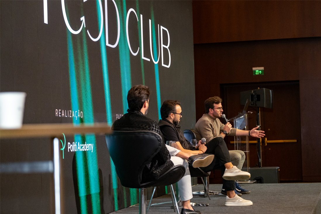 Três pessoas sentadas no palco em poltronas, participando de um painel de discussão em um evento chamado "FOOD CLUB", com microfones e uma tela grande atrás delas.