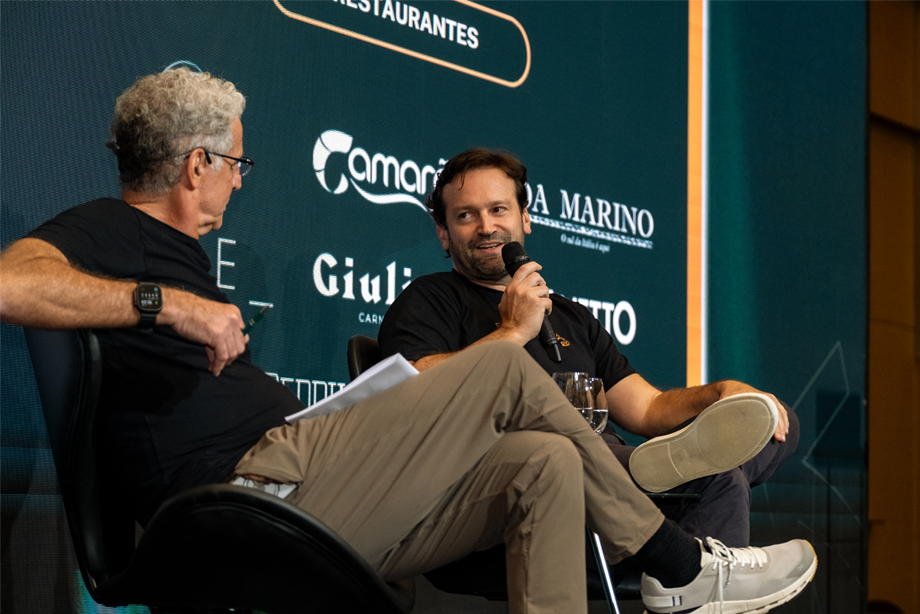 Dois homens estão sentados no palco conversando; um deles segura um microfone e sorri enquanto fala, com logotipos de restaurantes visíveis em uma tela atrás deles.