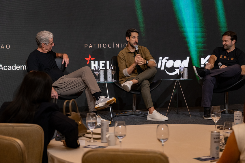 Três homens sentados no palco em um painel de discussão, cada um segurando um microfone, enquanto os membros da plateia assistem das mesas em primeiro plano.