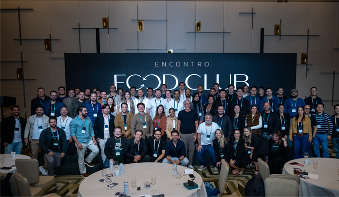 Um grande grupo de pessoas posa para uma foto em grupo em frente a uma placa onde se lê "Encontro Food Club" em uma sala de conferências.