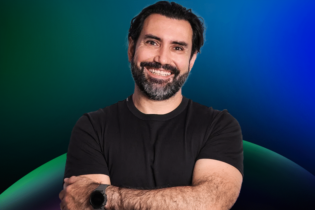Homem com cabelo e barba escuros, vestindo camiseta preta e smartwatch, sorri com os braços cruzados em um fundo gradiente azul e verde, representando a Lista de Espera do Acelera Food Nation de 2026.