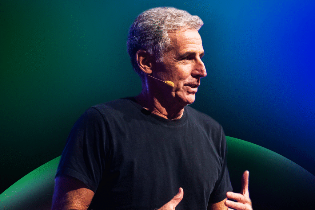 Um homem de cabelos grisalhos, vestindo uma camiseta preta e um microfone com fone de ouvido, gesticula enquanto fala sobre o Acelera Food Nation em um fundo gradiente azul e verde.