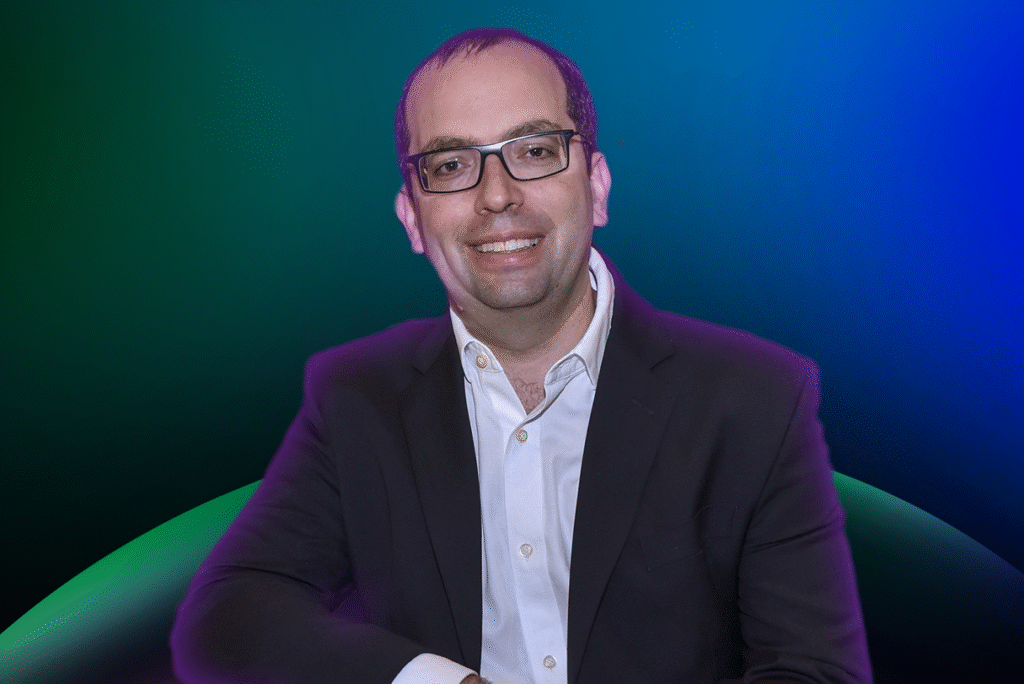 Um homem usando óculos, camisa branca e paletó escuro posa em um fundo gradiente azul e verde, representando a Lista de Espera de 2026 do Acelera Food Nation.