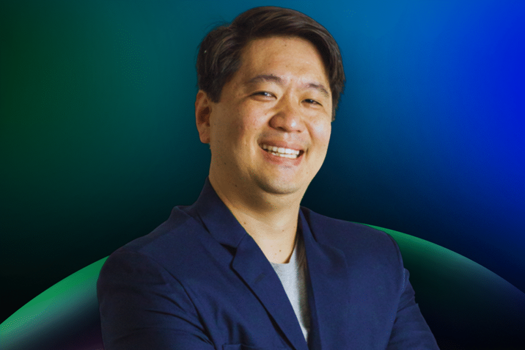 Um homem de cabelos escuros curtos, vestindo um blazer azul-marinho e uma camisa clara, sorri para a câmera contra um fundo gradiente azul e verde, representando o Acelera Food Nation enquanto aguardam ansiosamente a Lista de Espera de 2026.