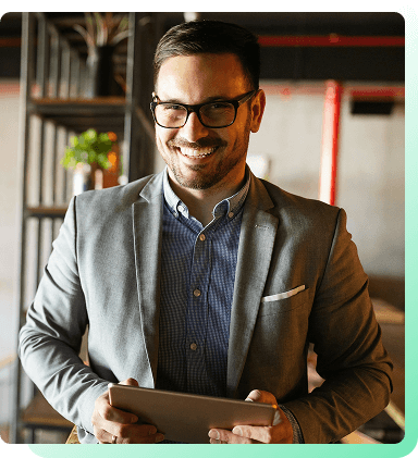 Homem usando óculos e blazer cinza sorri enquanto segura um tablet em um ambiente de escritório moderno. Prateleiras e uma planta são visíveis ao fundo.