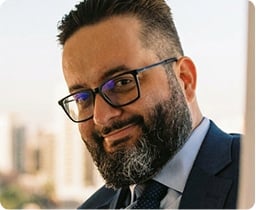 Homem com barba e óculos, vestindo terno e gravata, sorrindo levemente, com uma paisagem urbana desfocada ao fundo.