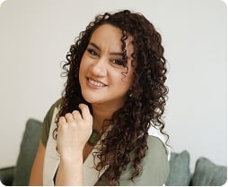 Mulher de cabelos castanhos encaracolados, vestindo um top verde e branco, senta-se em um sofá e sorri para a câmera com a mão apoiada no queixo.