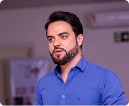 Um homem de cabelos escuros e barba, vestindo uma camisa azul, está em pé dentro de casa e parece estar falando.