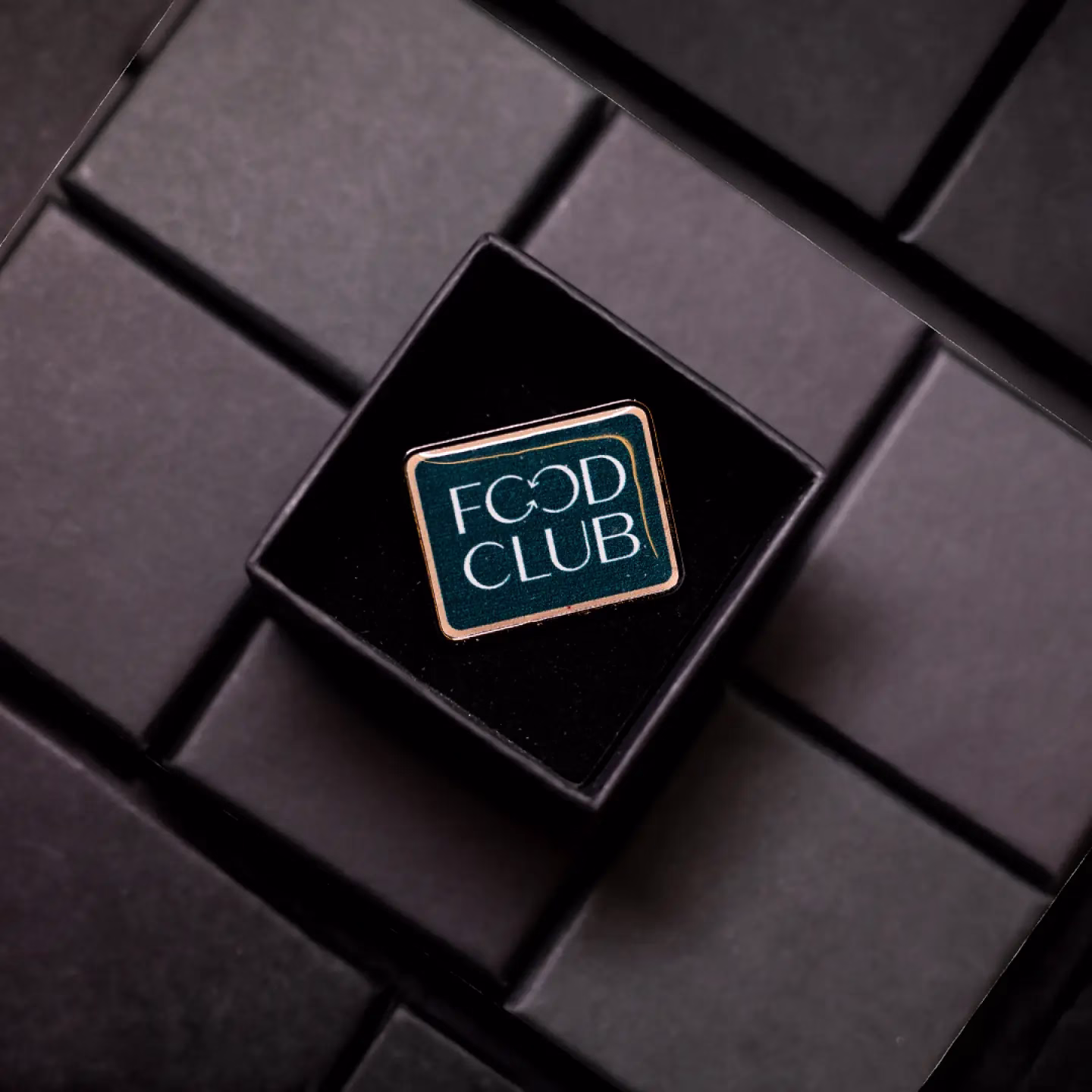 Um distintivo retangular com as palavras "FOOD CLUB" em branco sobre um fundo escuro, colocado em uma caixa preta em uma superfície preta com padrão de grade.